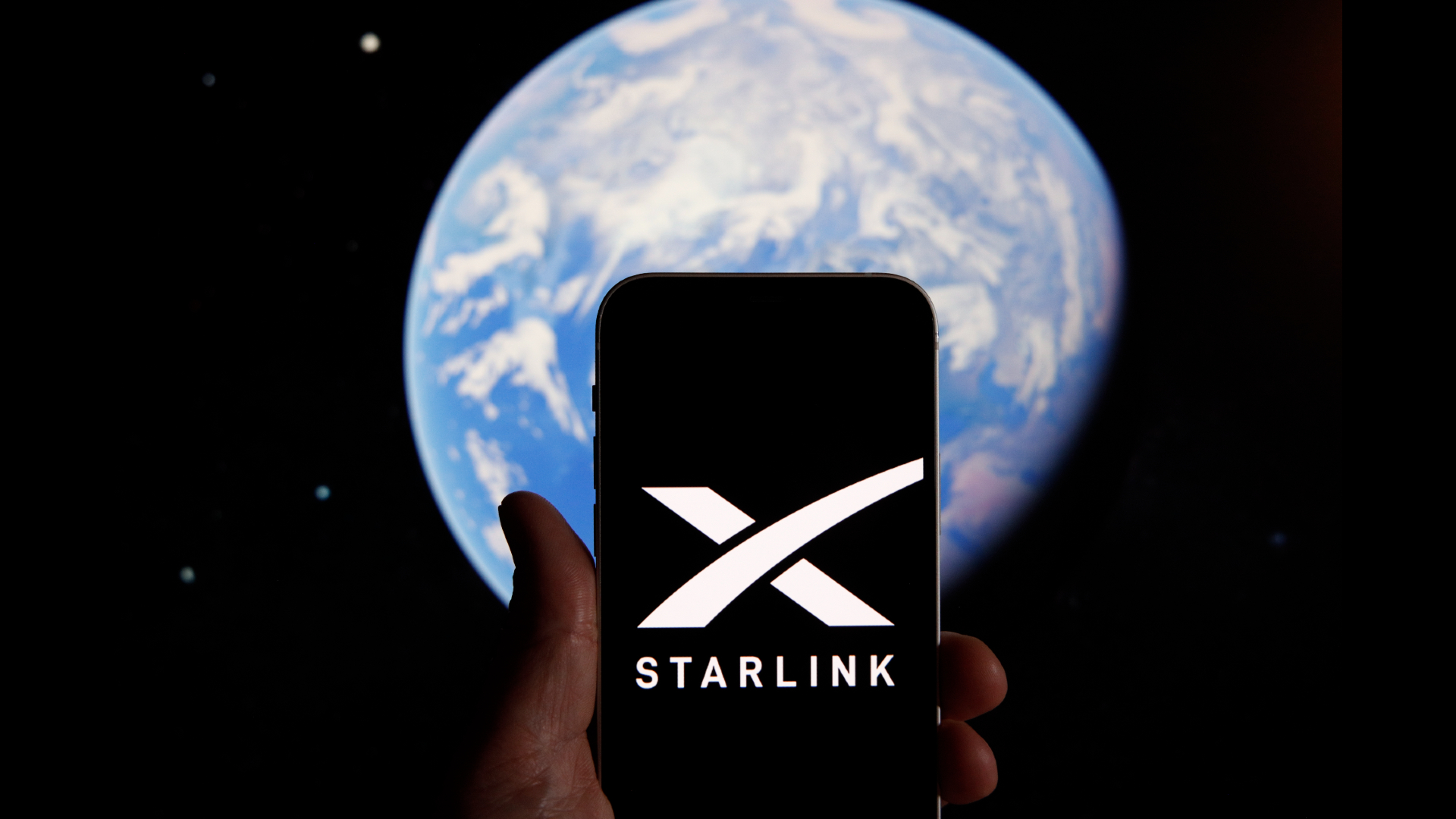 In the foreground is a hand holding a phone with the Starlink logo on the screen behind it in the background of the image is planet Earth.